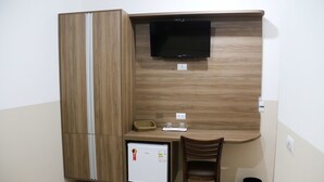 A compact room with a built-in desk, a flat-screen TV, a mini-fridge, and a trash bin. - Hotel Vale das Artes (Embu das Artes)