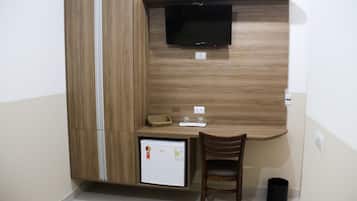 Una habitación compacta con escritorio integrado, televisión de pantalla plana, refrigerador pequeño y un cesto de basura.