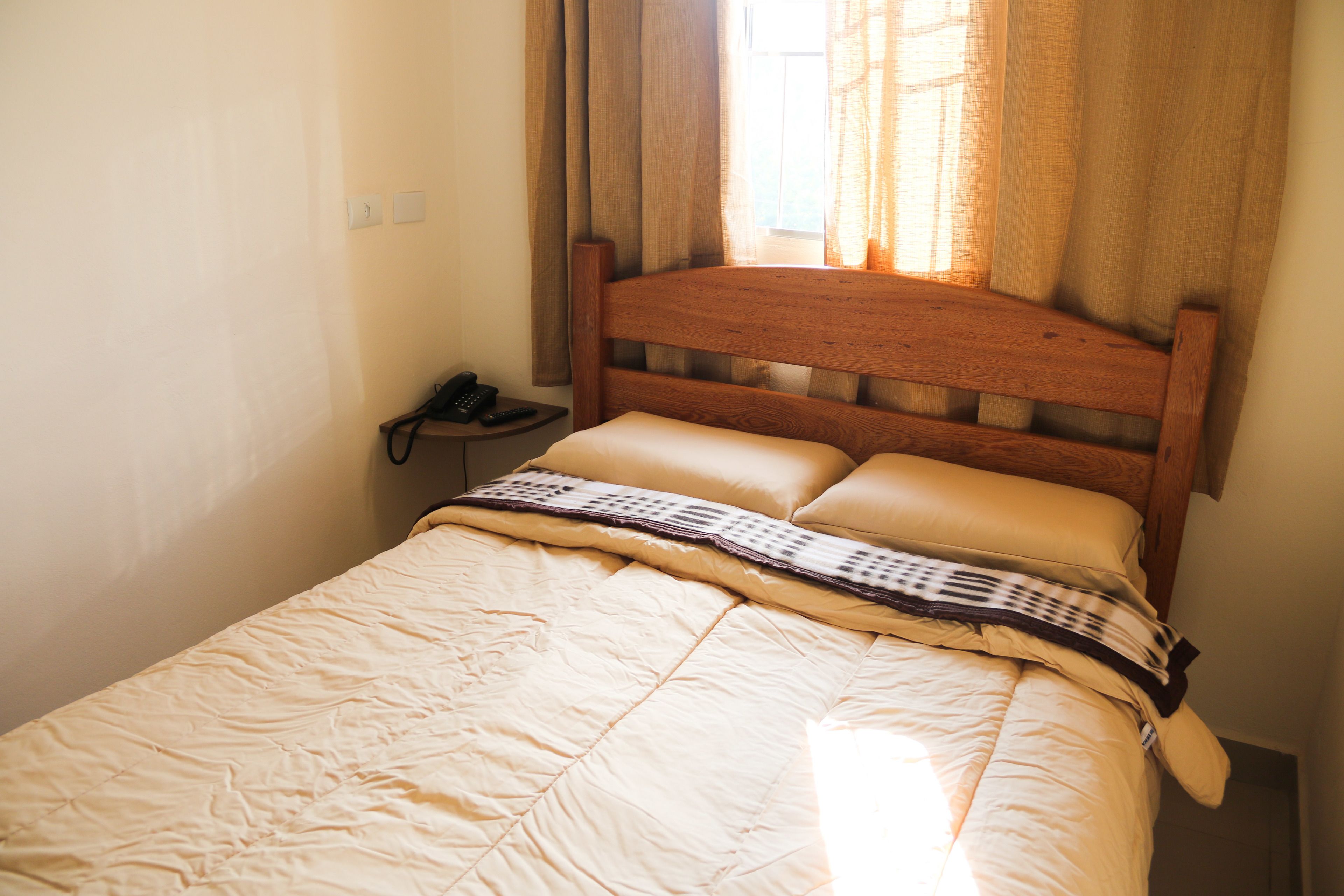 A neatly made bed with a wooden headboard, a bedside telephone, and a window with curtains.