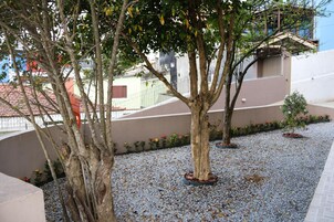 Um pátio com árvores, chão de cascalho e um edifício com parede marrom e telhado vermelho.