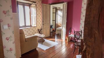 Een kamer met houten vloer, een beige bank, een kleine tafel en een stoel. Er is een raam met bloemenbehang en een deur die naar een andere kamer leidt.