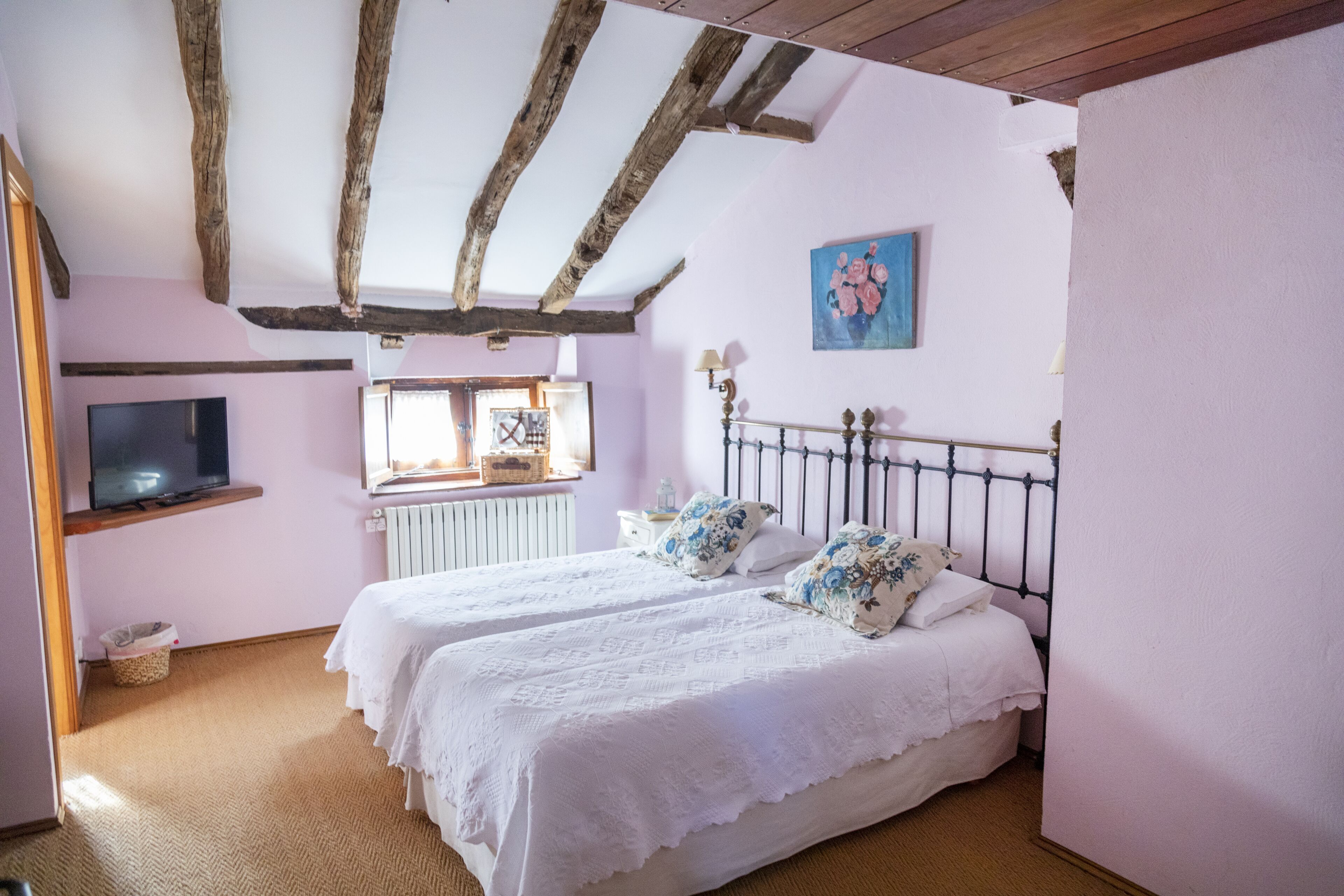 Un dormitorio con dos camas, vigas de madera en el techo, un televisor en la pared y una ventana con luz natural.