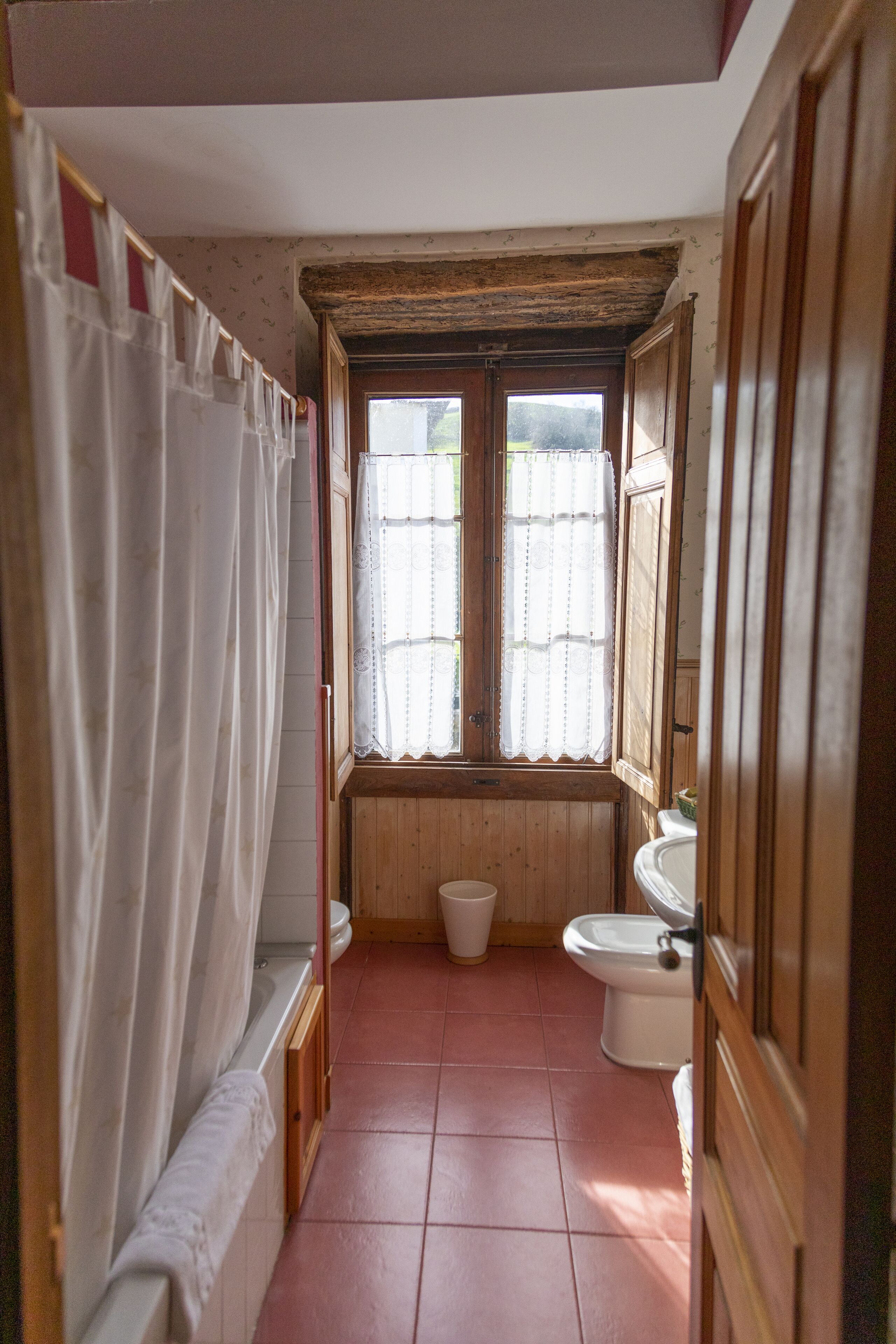A narrow bathroom with a bathtub, toilet, and sink. There is a window with a curtain, and the walls are wooden.