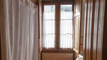 A narrow bathroom with a bathtub, toilet, and sink. There is a window with a curtain, and the walls are wooden.