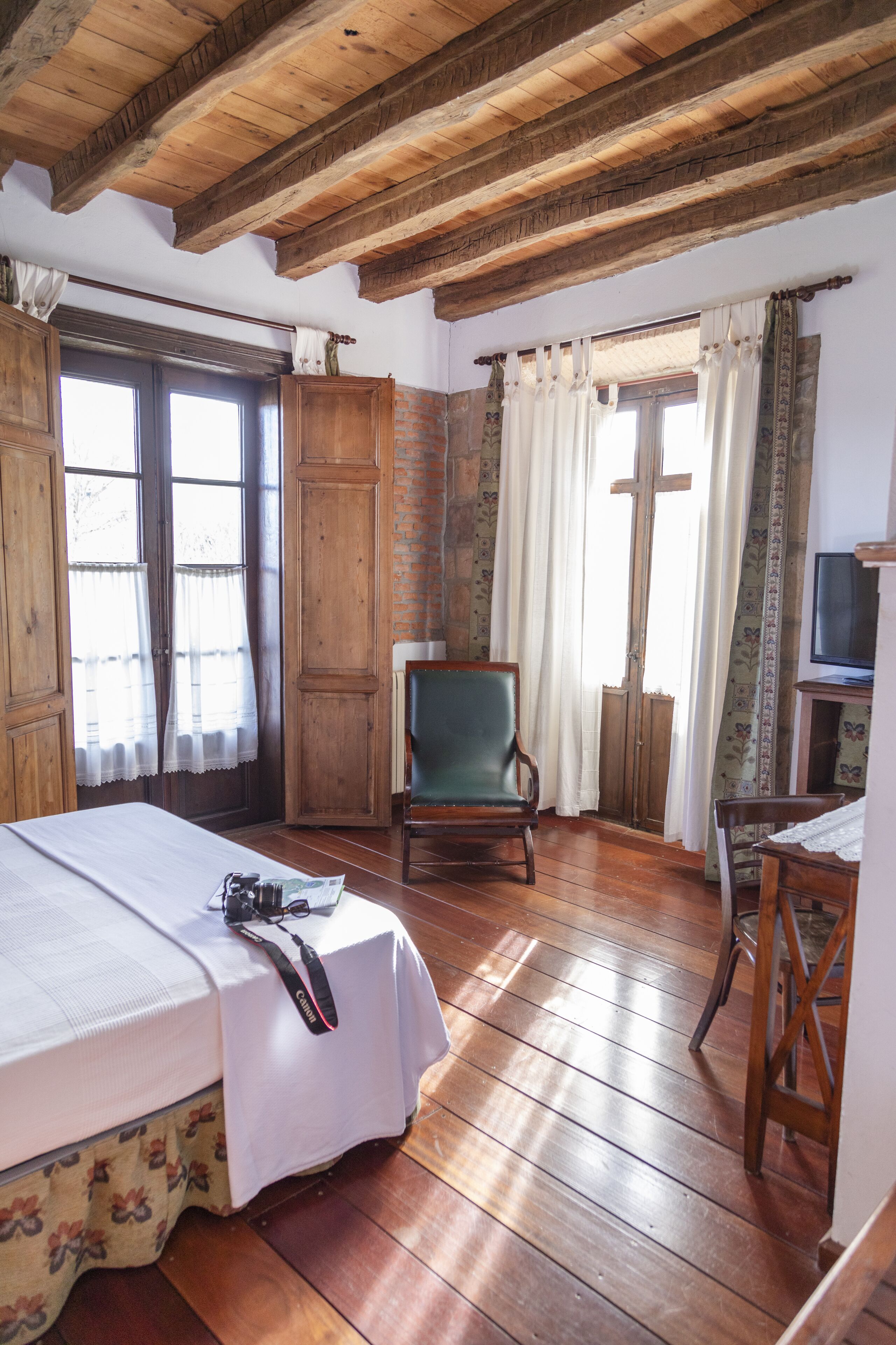 A room with wooden floors, a bed with a white bedspread, a green chair, a wooden table with a television, and a cross-shaped window.