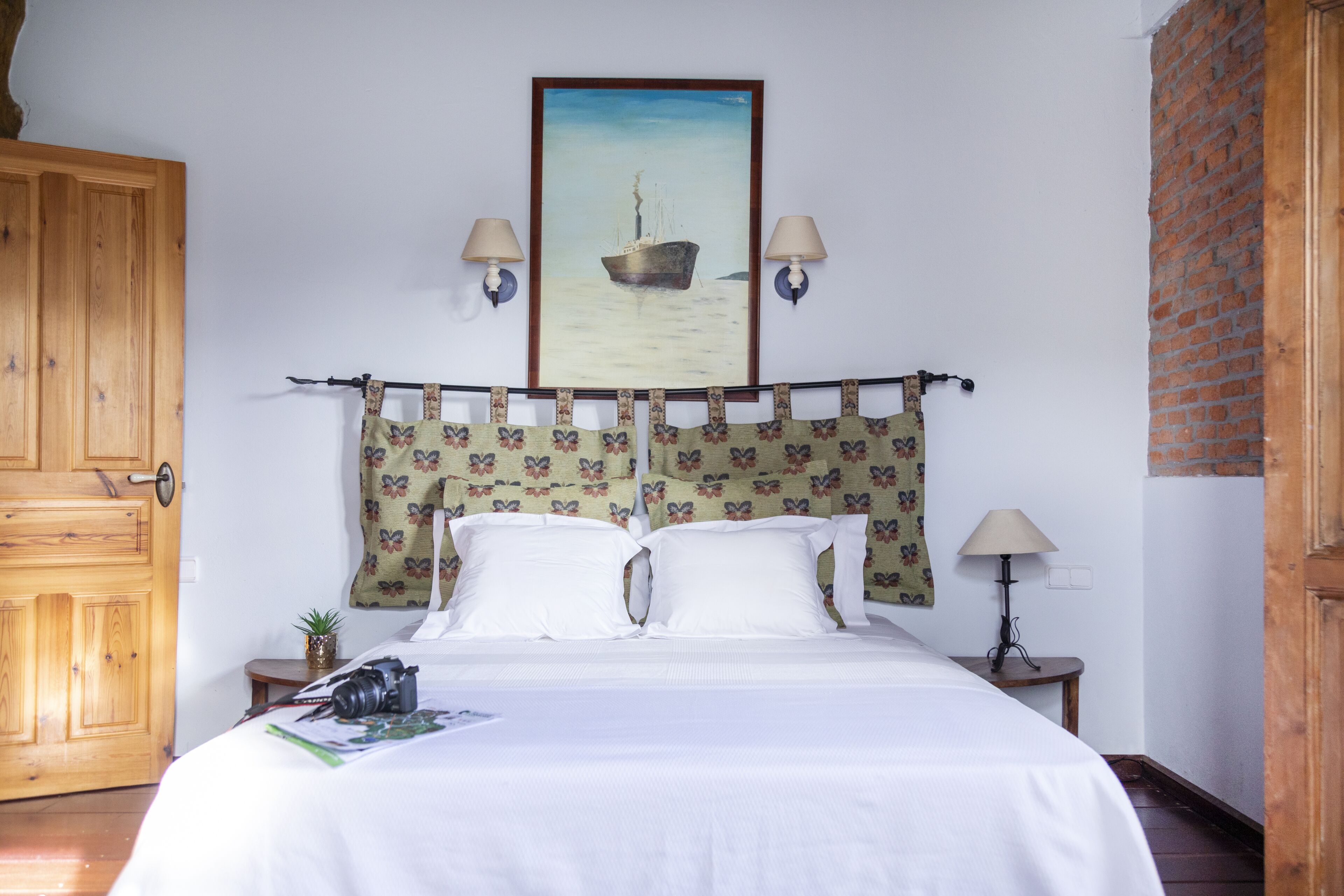 Un dormitorio con una cama, dos lámparas de pared, un cuadro de un barco, una puerta de madera y una pequeña mesa con una lámpara.