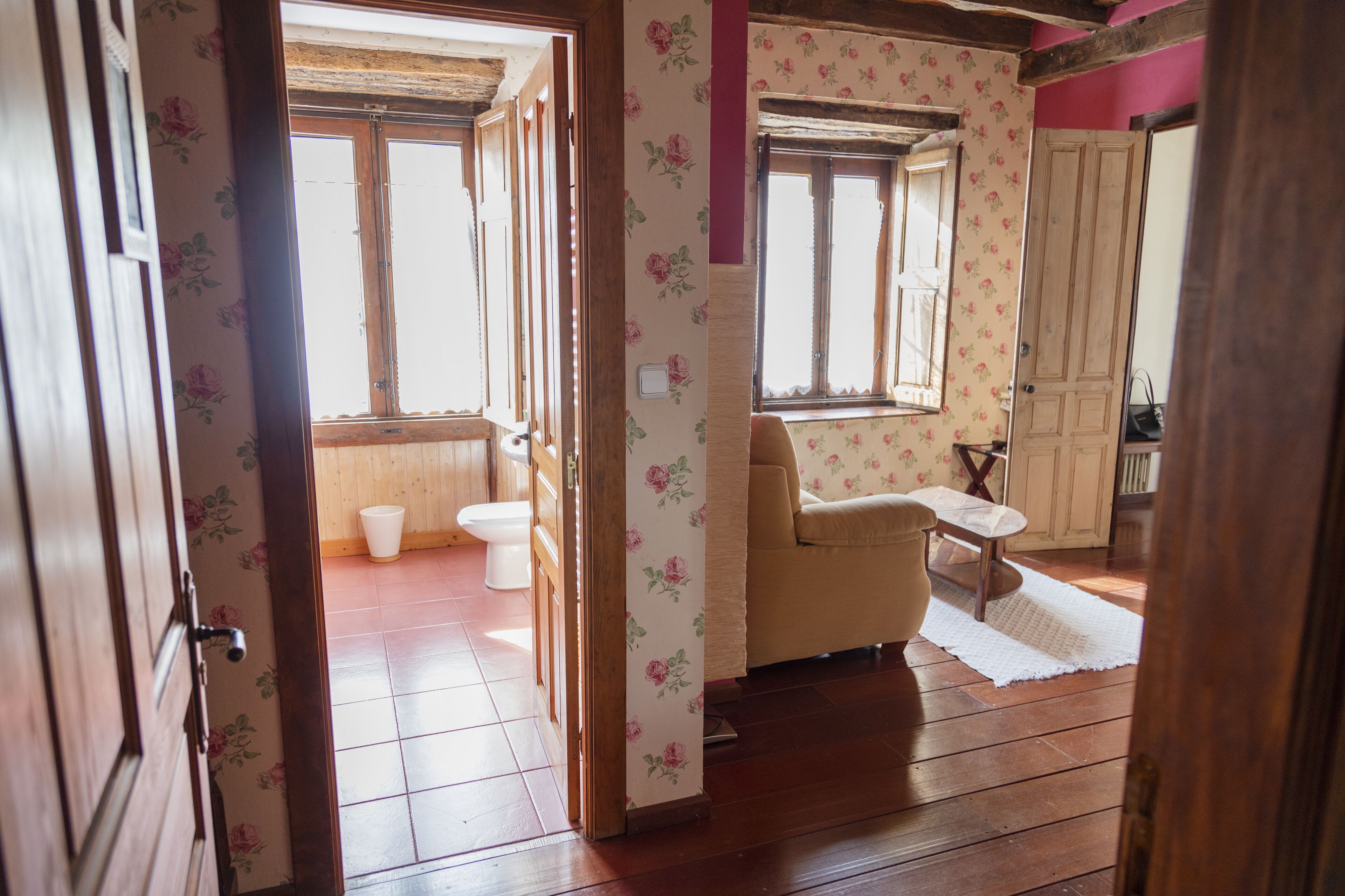 Una habitación con suelo de madera, un sofá color beige, una mesita y un ventanal con papel tapiz floral.