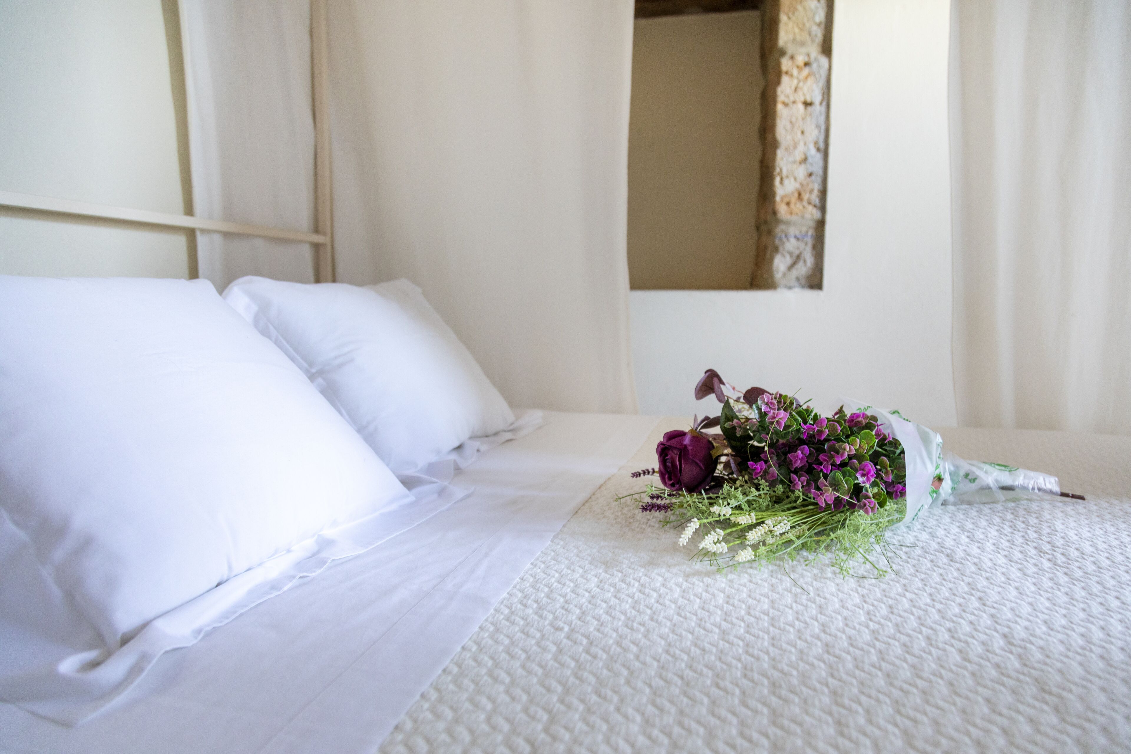 Una cama impecable con sábanas blancas y un ramo de flores sobre un edredón blanco texturizado.