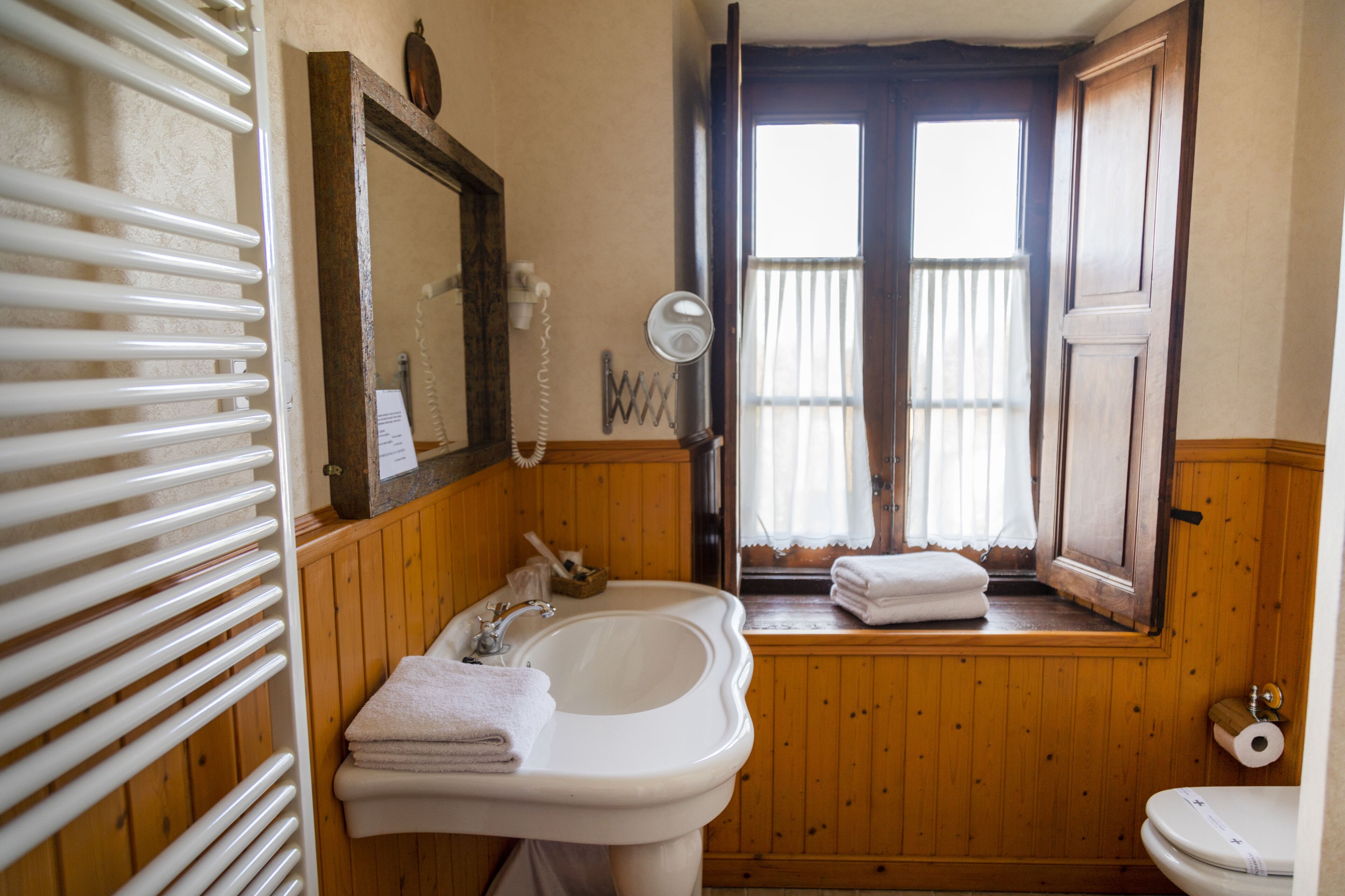 Une salle de bain avec des lambris en bois, un lavabo, un miroir, des toilettes et une fenêtre avec des rideaux.