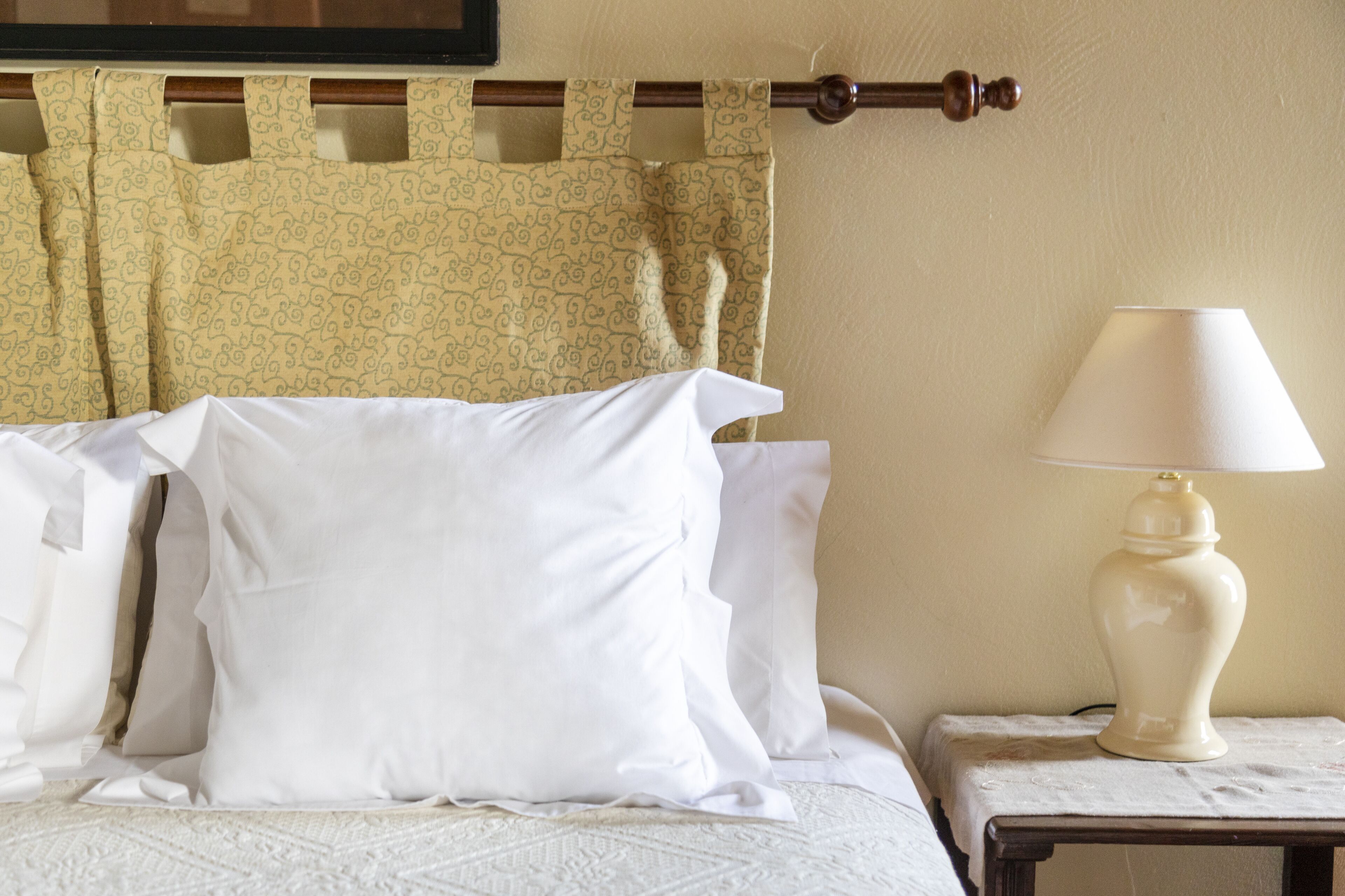A neatly made bed with white linens and pillows, a bedside table with a lamp, and a patterned curtain.