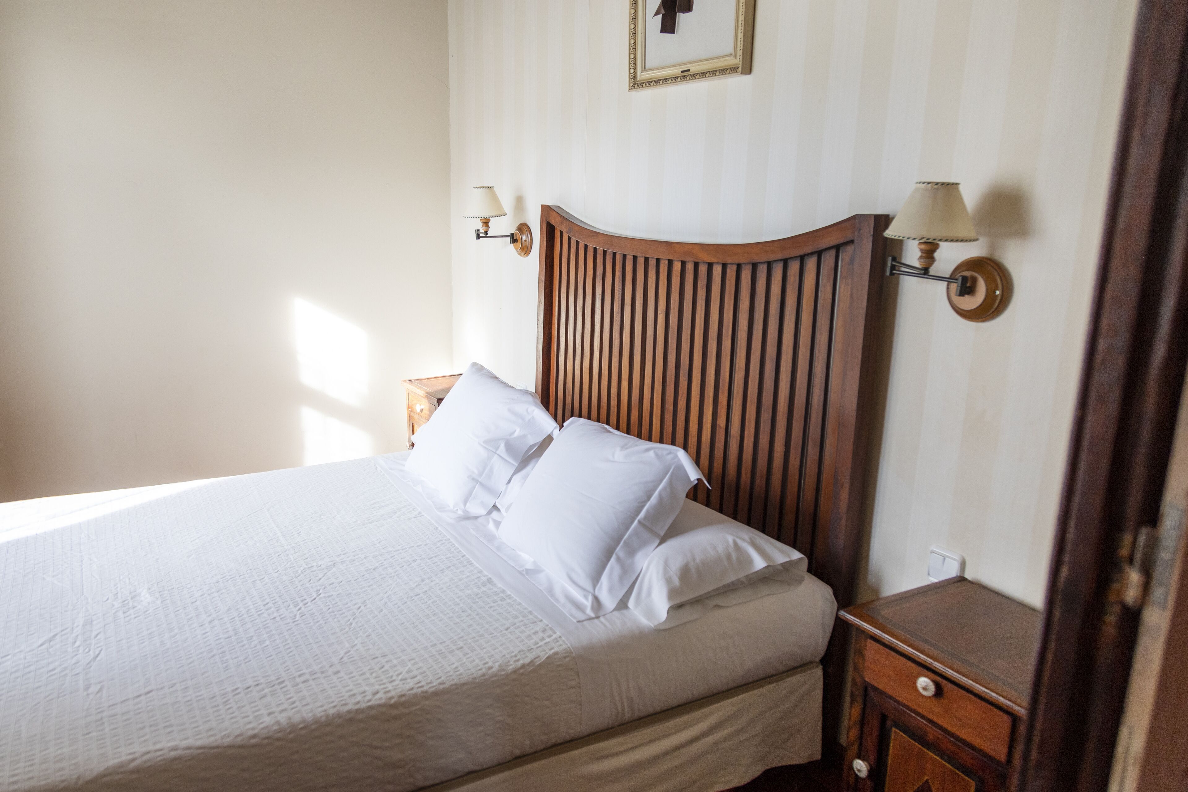 Una cama impecablemente hecha con sábanas blancas y cabecero de madera, con lámparas fijadas a la pared a ambos lados.