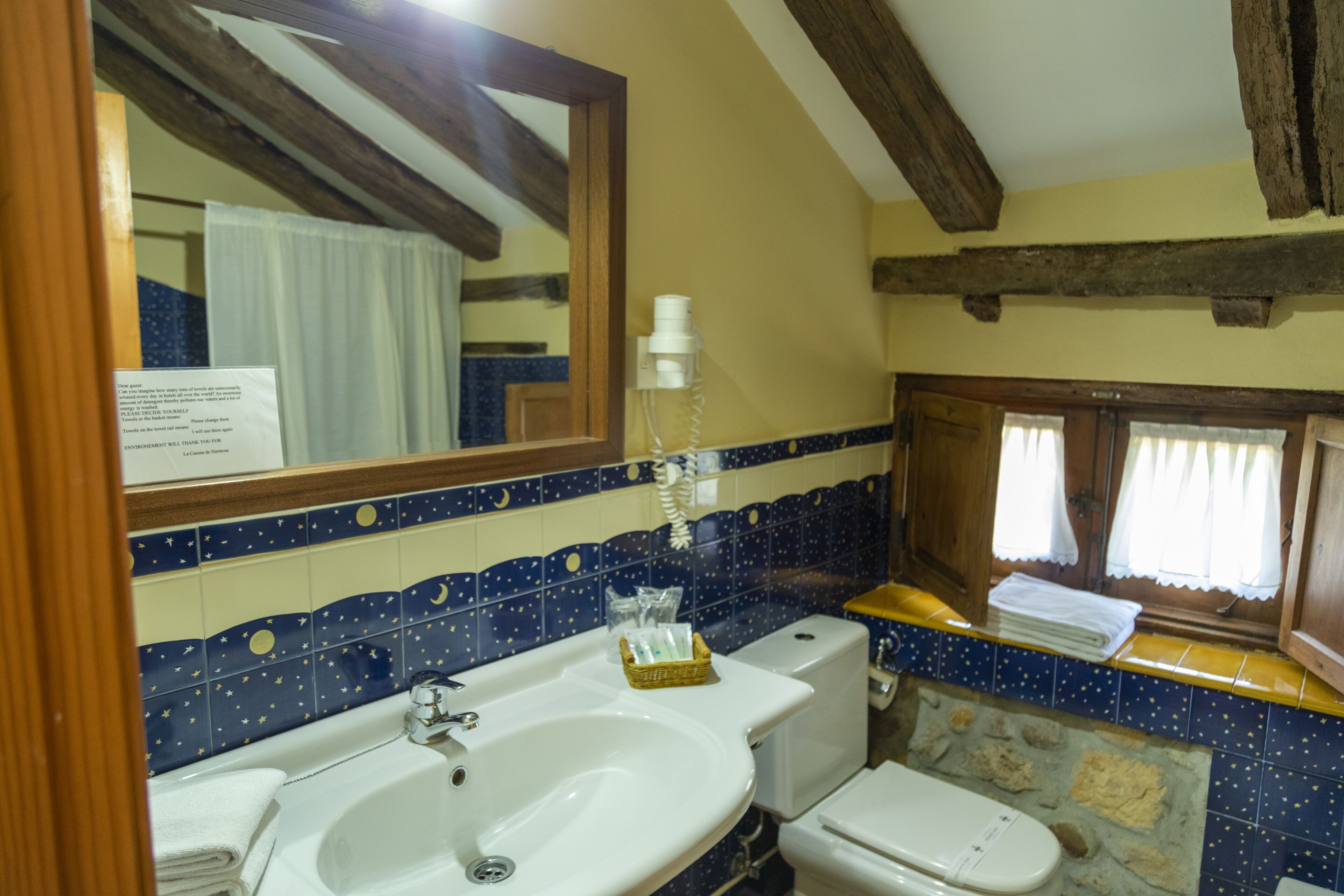 Un baño con lavamanos, inodoro y espejo. Hay una ventana con cortinas y un perchero con toallas.