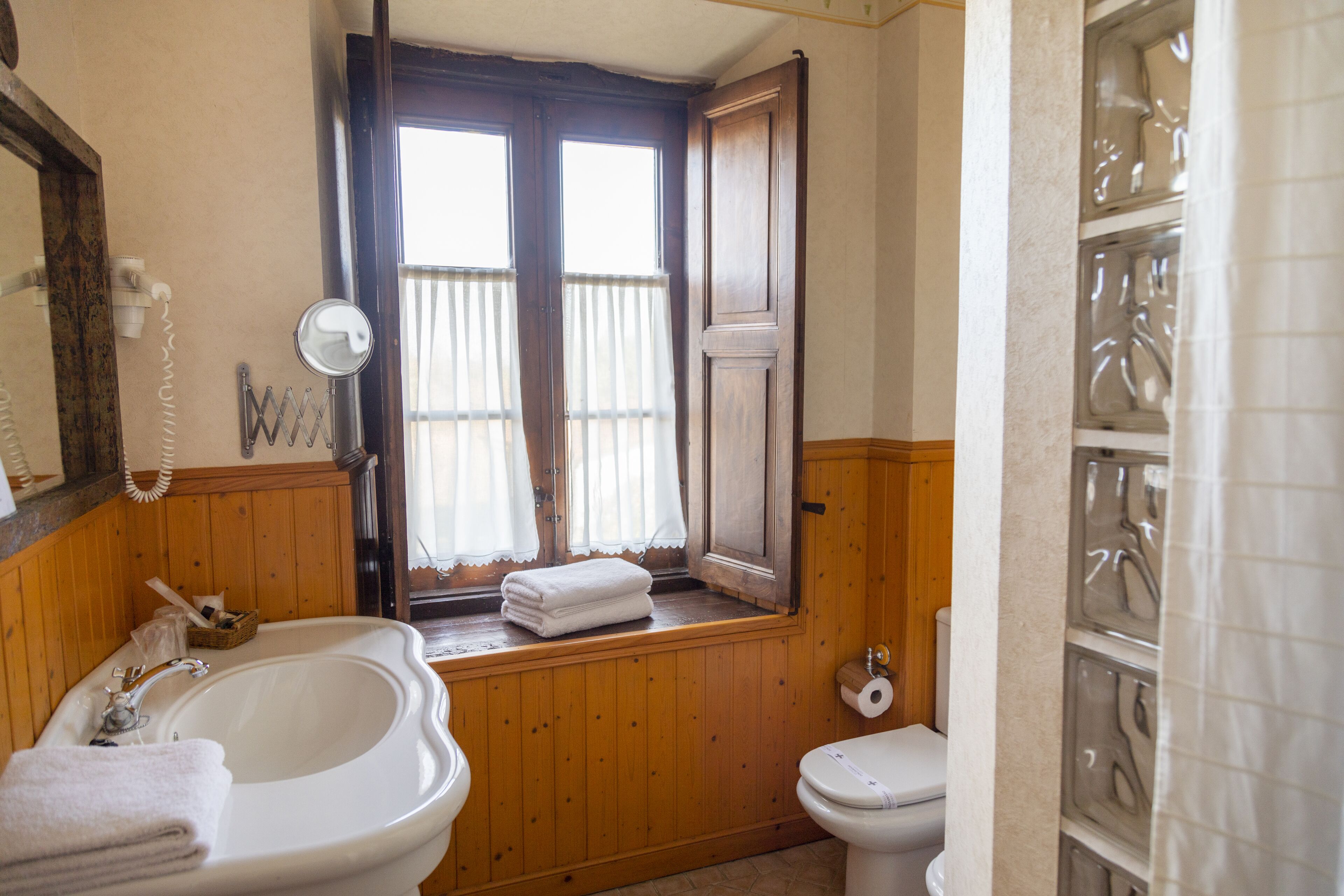 Un baño con tocador de madera, lavamanos, inodoro y ventana con cortinas.
