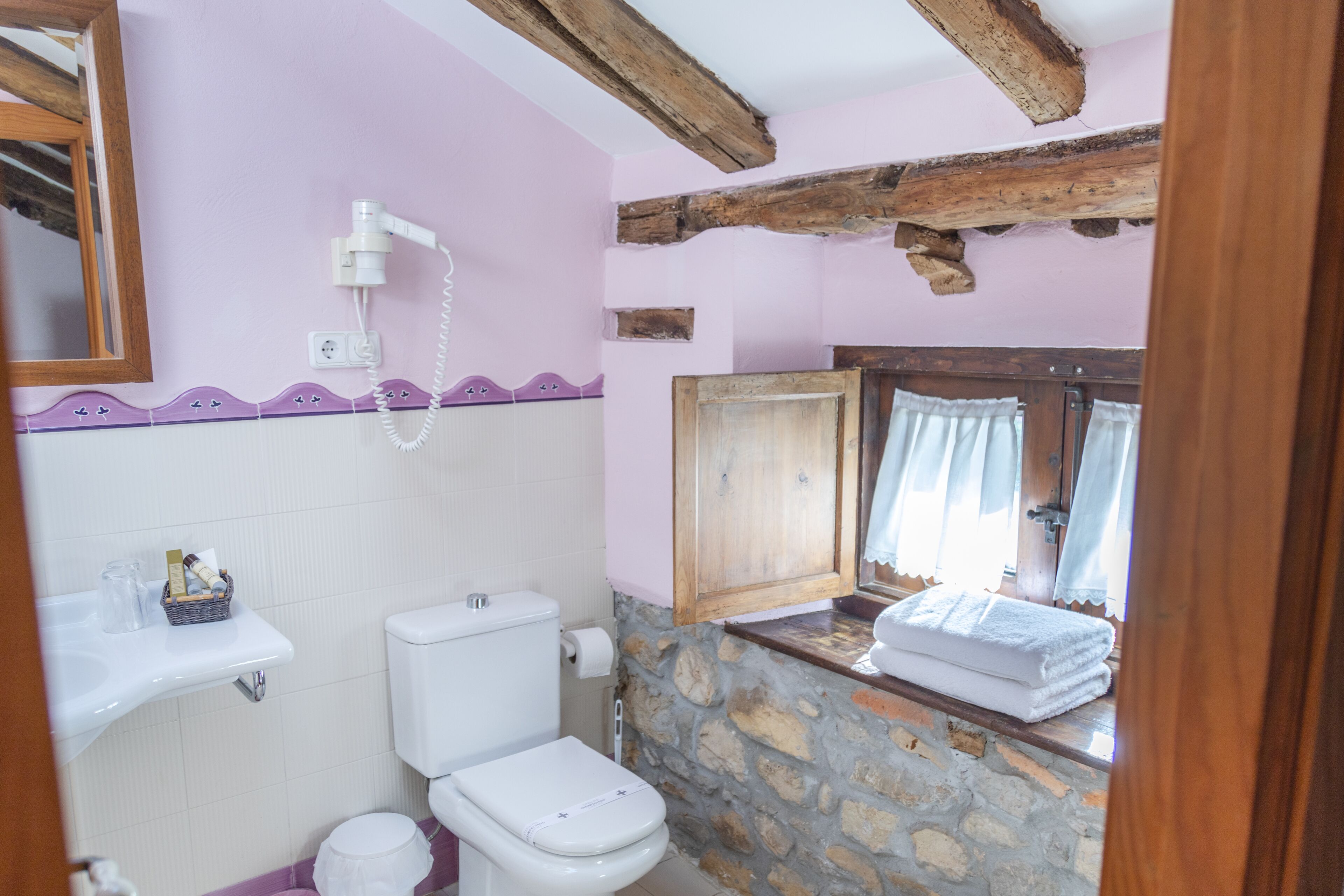 Un baño con pared de piedra, vigas de madera, inodoro blanco, lavamanos y una ventana con cortinas.