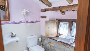 Un baño con pared de piedra, vigas de madera, inodoro blanco, lavamanos y una ventana con cortinas.