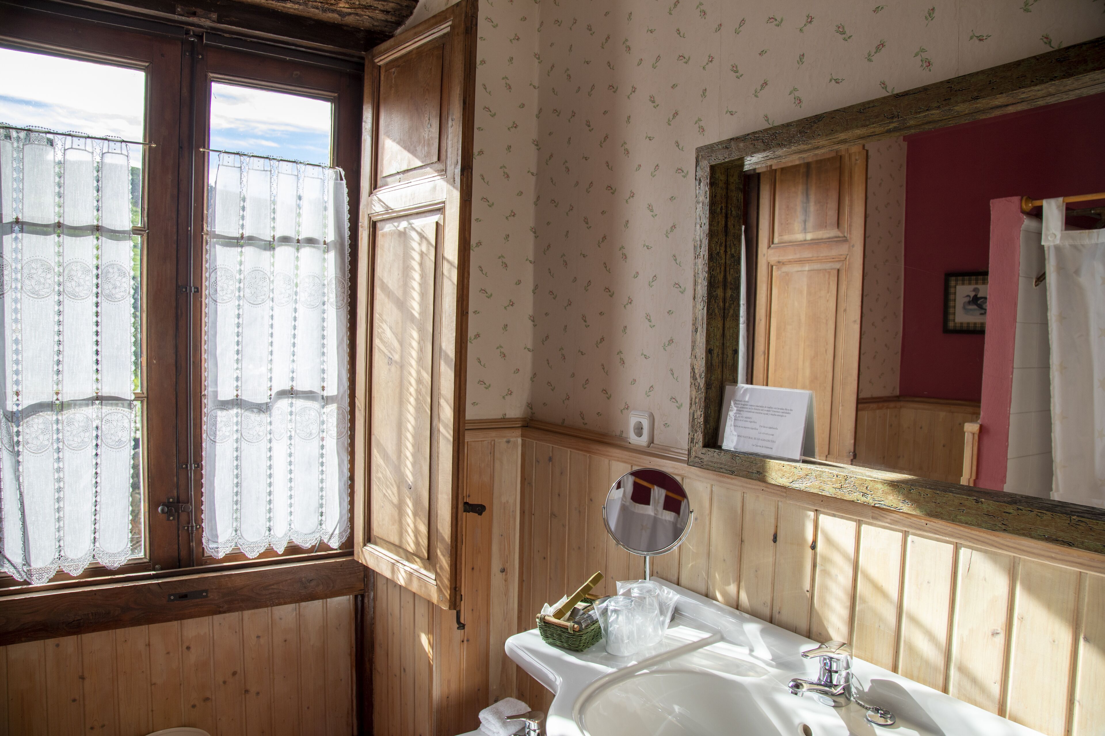 Un baño con paneles de madera, un espejo grande, un lavamanos y una ventana con cortinas de encaje.