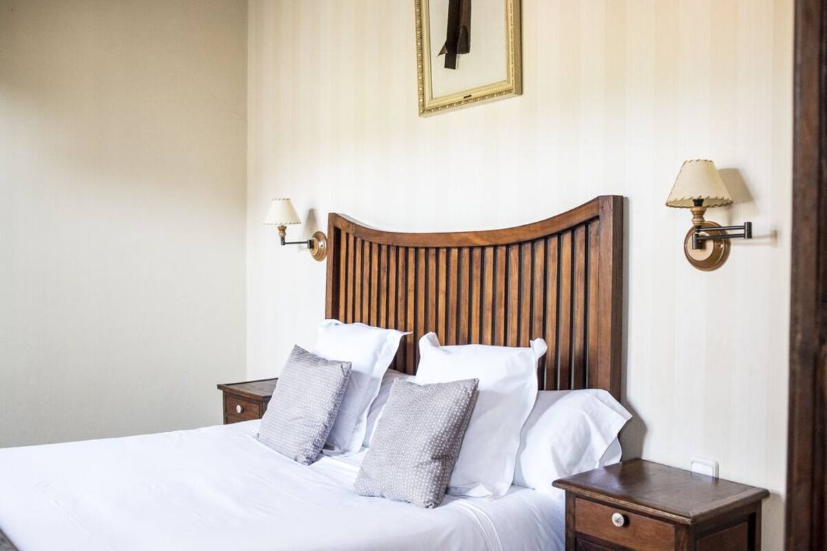 Una cama bien tendida con sábanas blancas y almohadas grises, un cabecero de madera y dos mesitas de noche con lámparas.