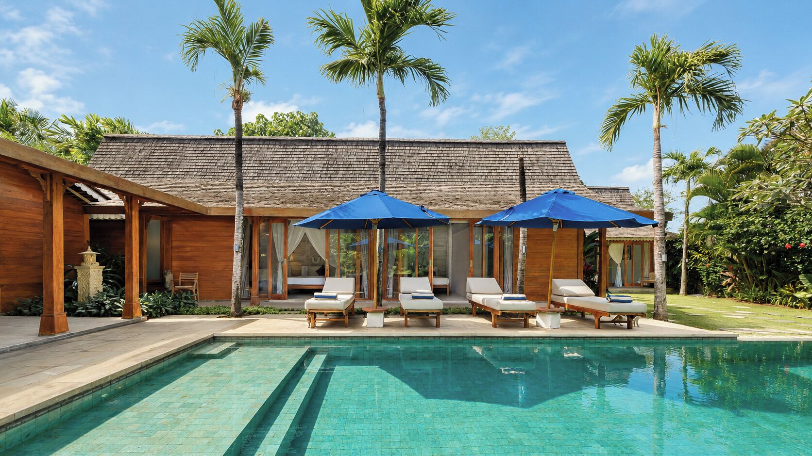 A poolside area with lounge chairs and umbrellas, a wooden building with a tiled roof, and palm trees.
