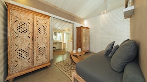 A room with a sofa, a wooden cabinet with intricate carvings, a white brick wall, and a doorway leading to another room. - Villa Little Mannao by Villa Finder (Kerobokan)