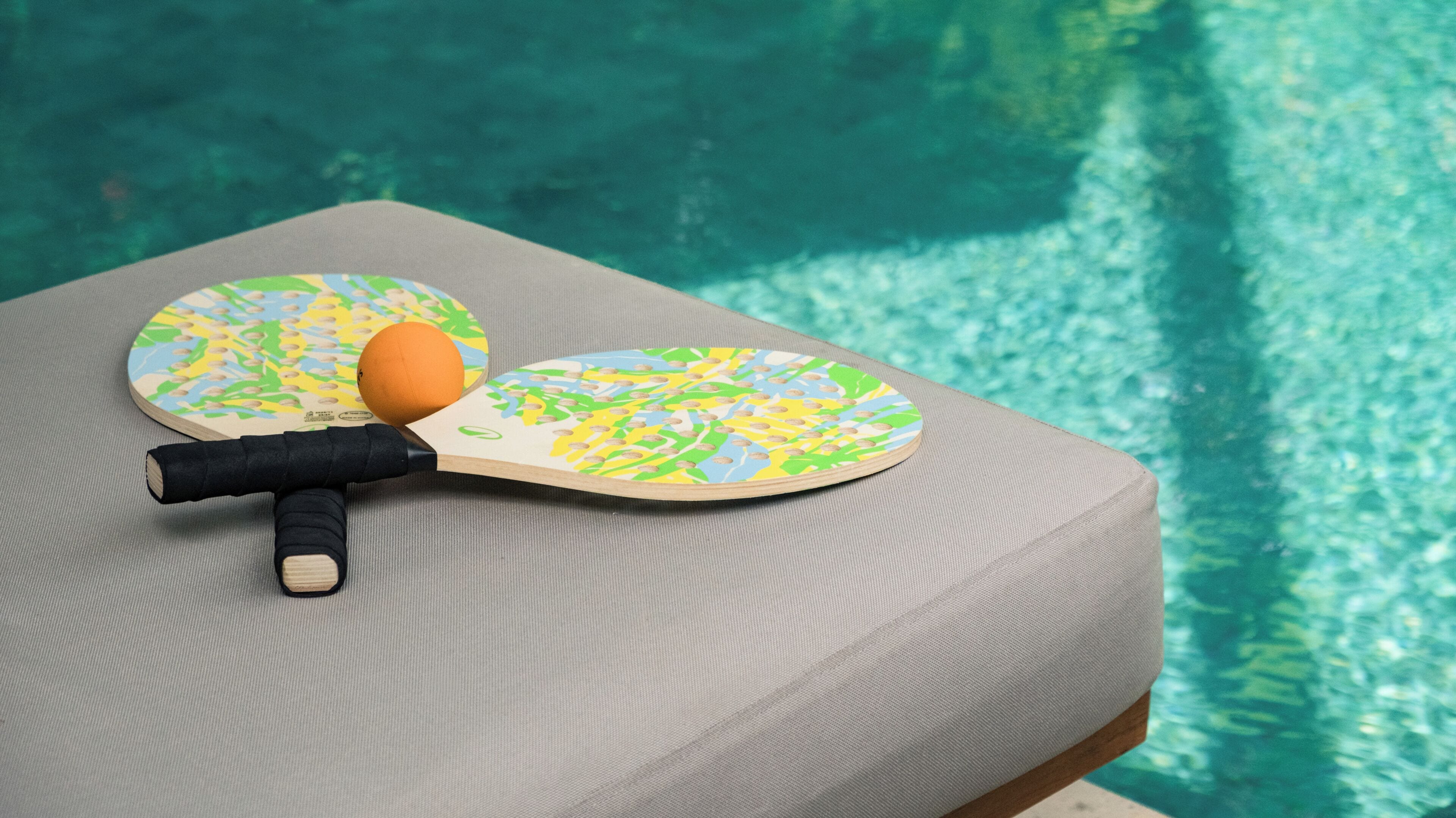 Two ping pong paddles and a ball on a cushioned surface by a pool.