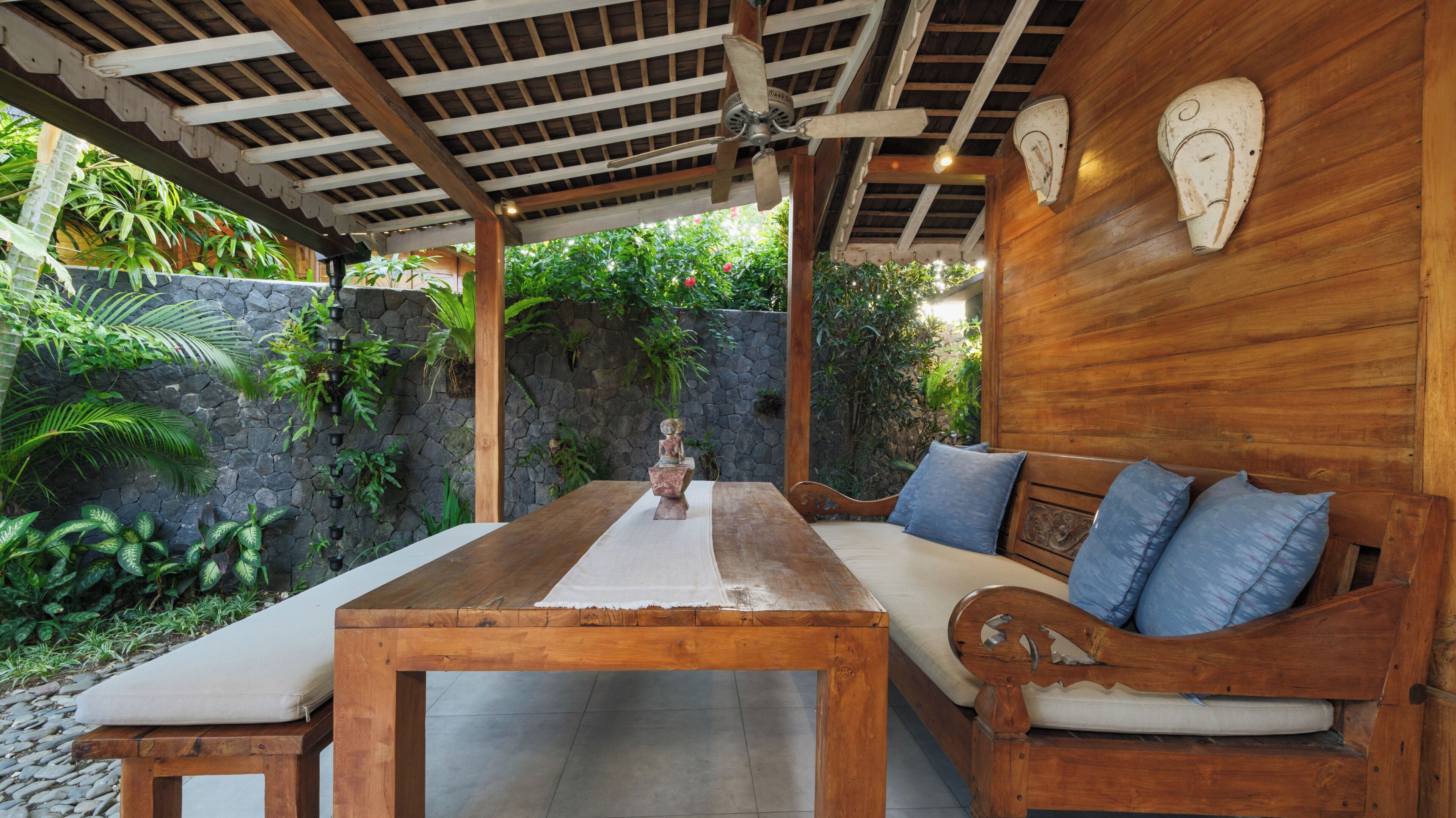 An outdoor seating area with a wooden table and bench, surrounded by greenery and a stone wall.