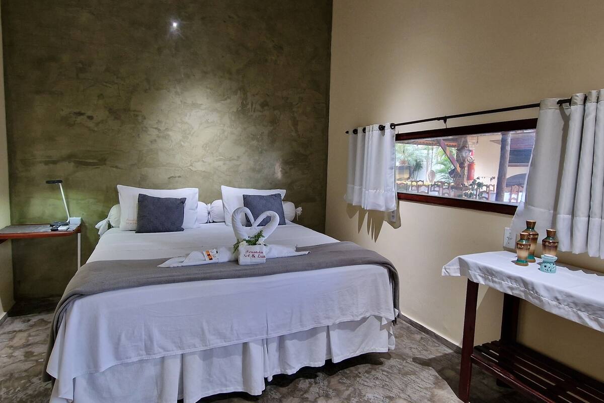 Ein ordentlich eingerichtetes Hotelzimmer mit einem großen Bett, einem Nachttisch mit Pflegeartikeln und einem Fenster mit Vorhängen.
