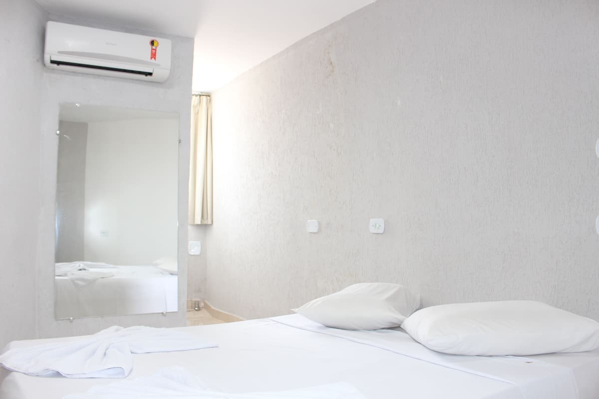 A hotel room with a bed, a mirror, an air conditioning unit, and a light switch on the wall.