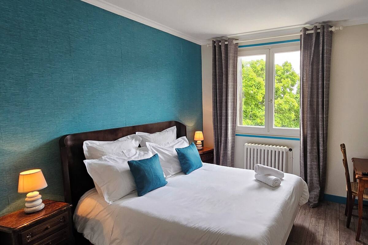 Quarto de hotel com cama, escrivaninha, cadeira, janela com cortinas, radiador e um quadro na parede.