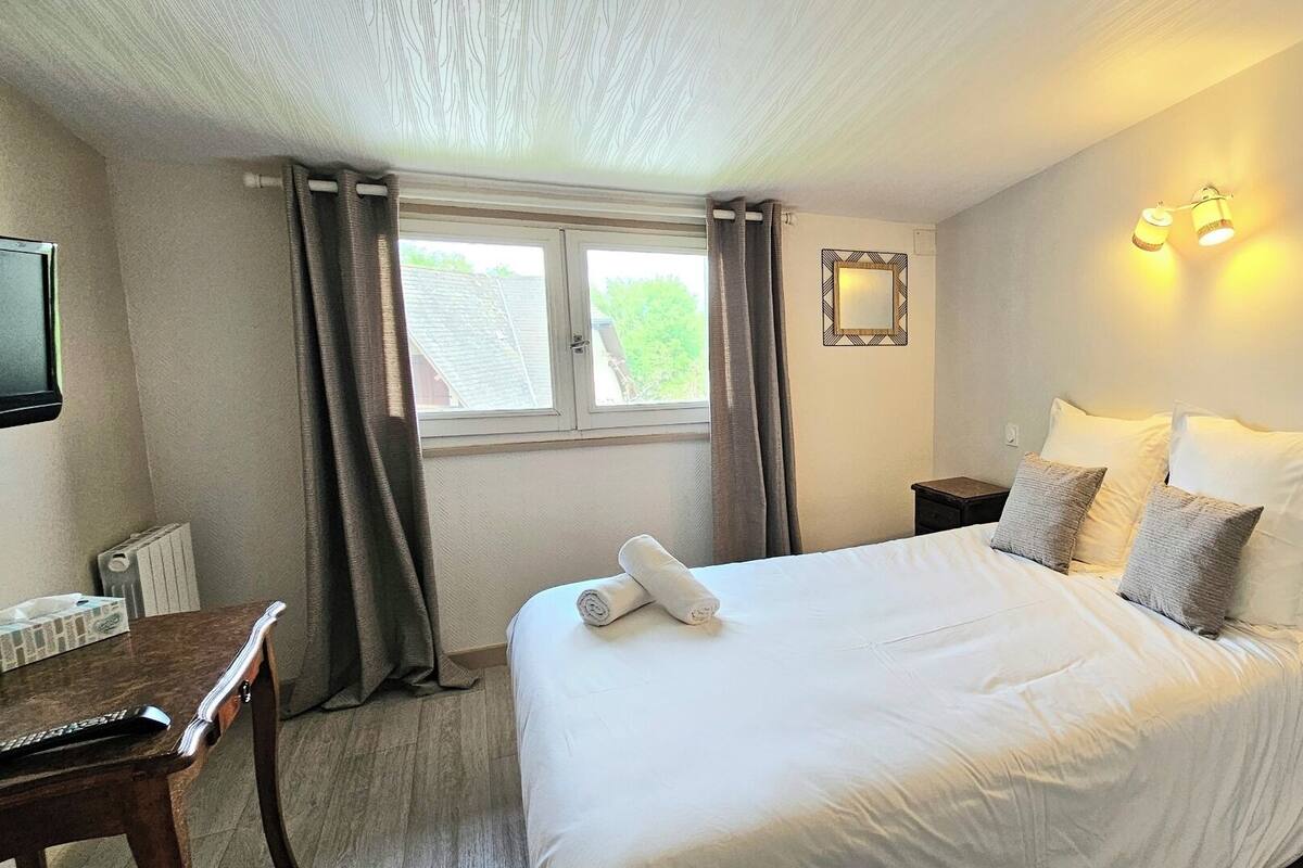 Cama bem arrumada com almofadas, mesa de cabeceira, abajur e janela com cortinas.