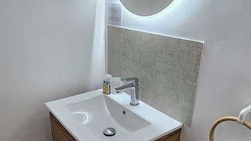 Ein modernes Badezimmer mit weißem Waschtisch, einem runden Spiegel mit integrierter Beleuchtung und einem Holzschrank.