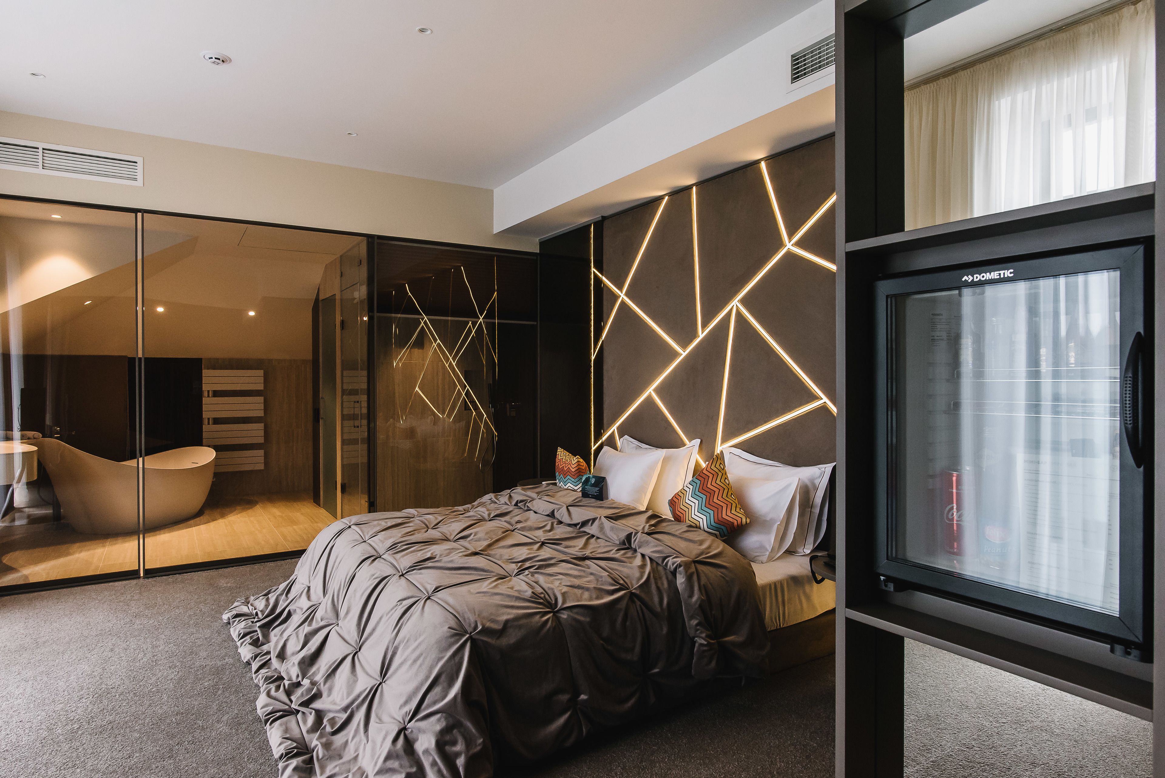 A modern bedroom with a large bed, a glass partition, a built-in TV, and a unique geometric light design on the wall.