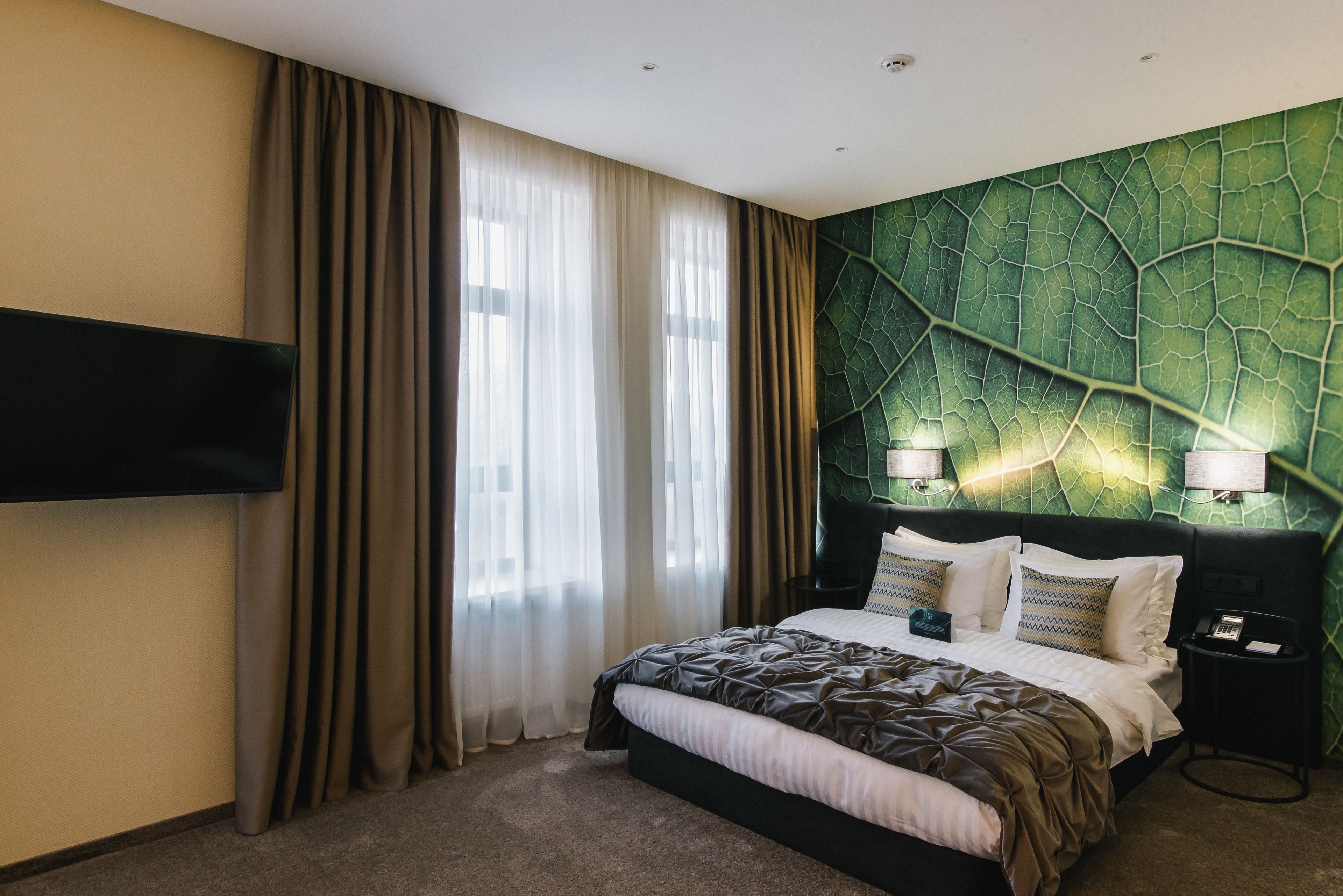 A hotel room with a bed, curtains, a flat-screen TV, and a green leaf-patterned wall.