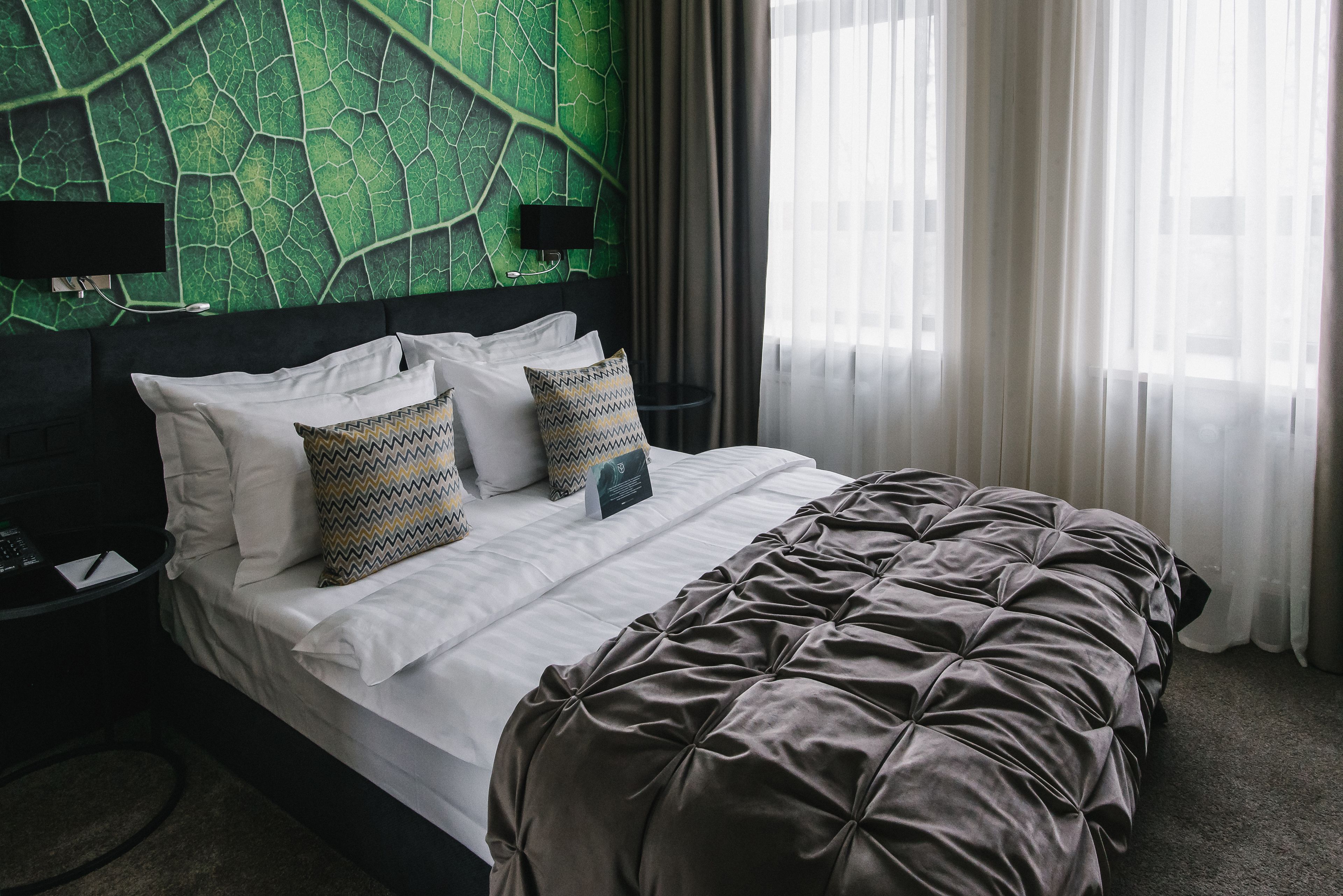 A neatly made bed with white linens and a brown tufted bedspread, a headboard with a green leaf-patterned wall, and a bedside table with a lamp.