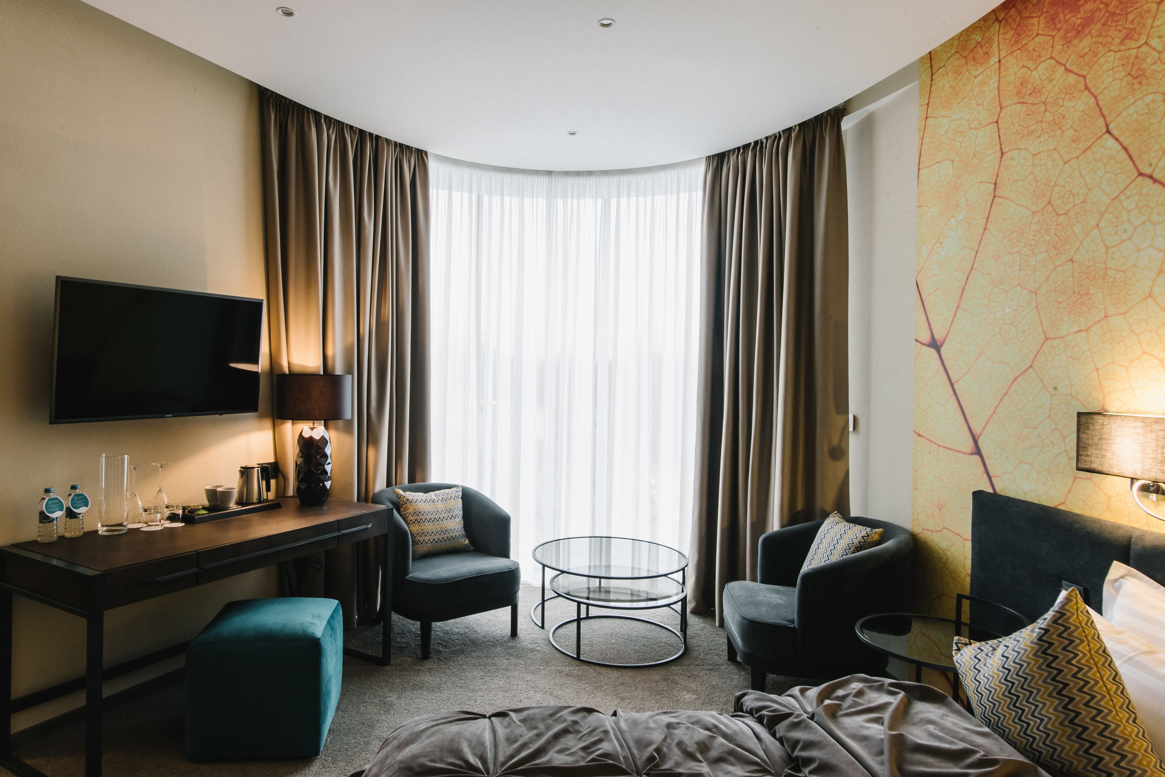 Ein Hotelzimmer mit Flachbildfernseher, Schreibtisch mit Lampe, zwei Sesseln, rundem Couchtisch und einem großen Fenster mit Vorhängen.