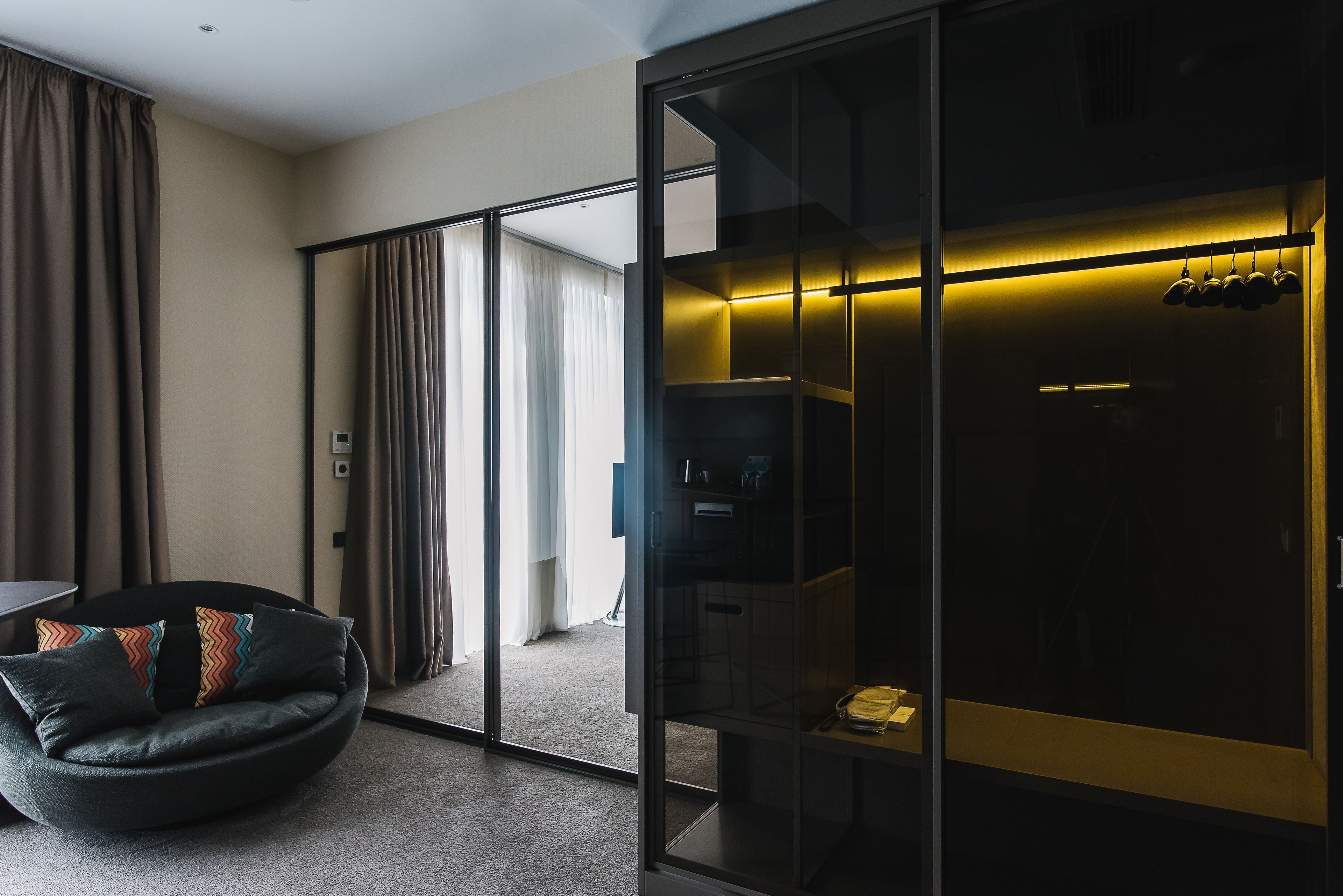 A modern hotel room with a glass partition, a black sofa with colorful cushions, and a visible kitchen area with hanging pots.