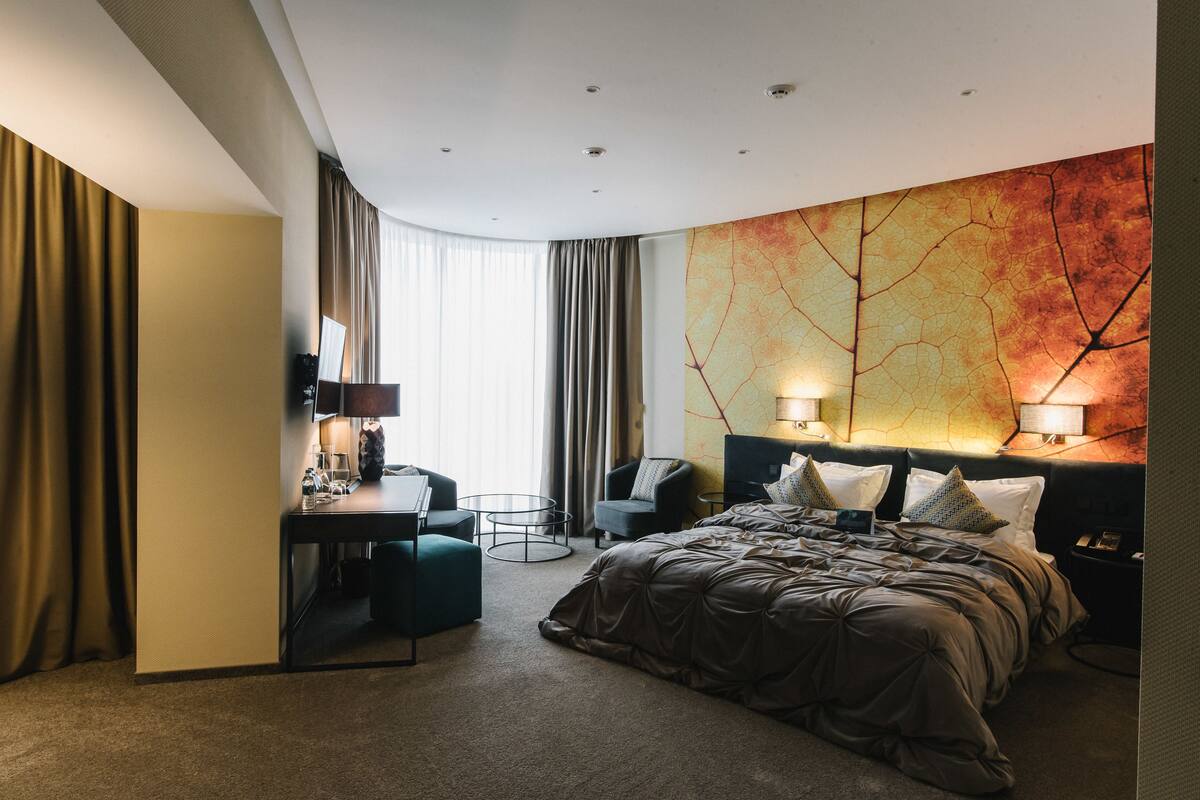 A modern hotel room with a large bed, a desk with a chair, a small round table, and a large abstract wall mural.
