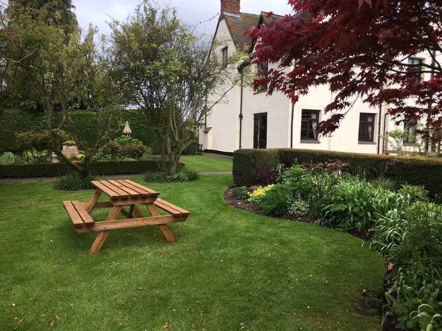 Uma mesa de piquenique de madeira num jardim bem cuidado, com um edifício branco ao fundo.