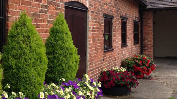 A brick building with a dark door, two windows, and a flower bed with red and purple flowers. - Marsh Farm (Rugeley)