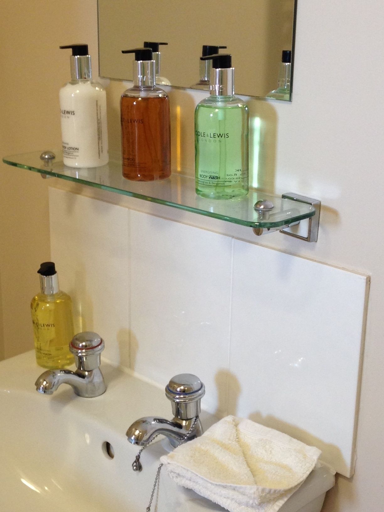 Ein Badezimmer mit einer Glasablage für Toilettenartikel, einem Waschbecken mit zwei Wasserhähnen und einem Handtuch auf dem Boden.