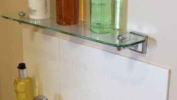 Ein Badezimmer mit einer Glasablage für Toilettenartikel, einem Waschbecken mit zwei Wasserhähnen und einem Handtuch auf dem Boden.
