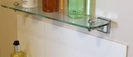 Ein Badezimmer mit einer Glasablage für Toilettenartikel, einem Waschbecken mit zwei Wasserhähnen und einem Handtuch auf dem Boden.