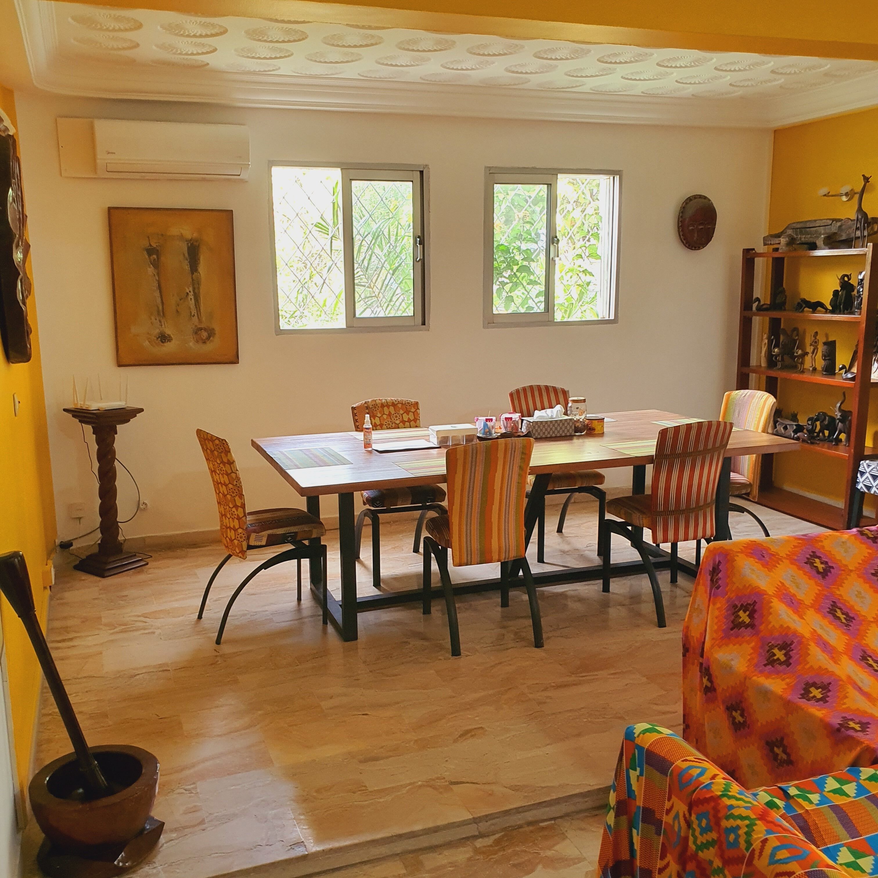 A dining area with a wooden table and chairs, a bookshelf, and a window with a view of greenery.