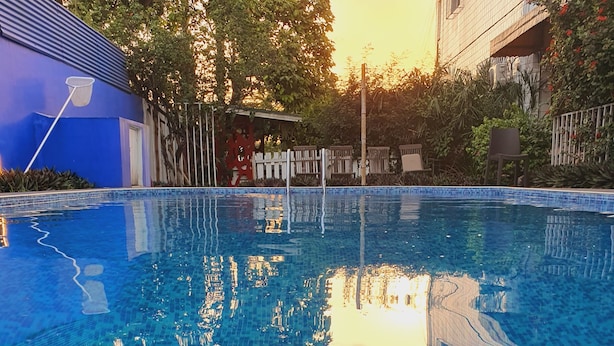 A clear swimming pool with a tiled edge, surrounded by greenery and a building with a blue facade. - Villa Mango (Abidjan)