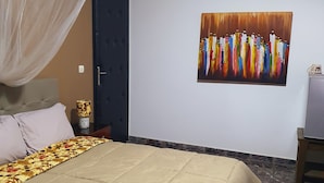 A bedroom with a bed, a nightstand, a decorative lamp, a door, and a painting on the wall. - Villa Mango (Abidjan)
