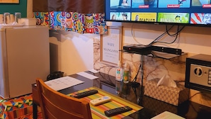 A hotel room with a television displaying a streaming service interface, a desk with a remote control, a chair, a small table, a tissue box, and a wall-mounted sign. - Villa Mango (Abidjan)