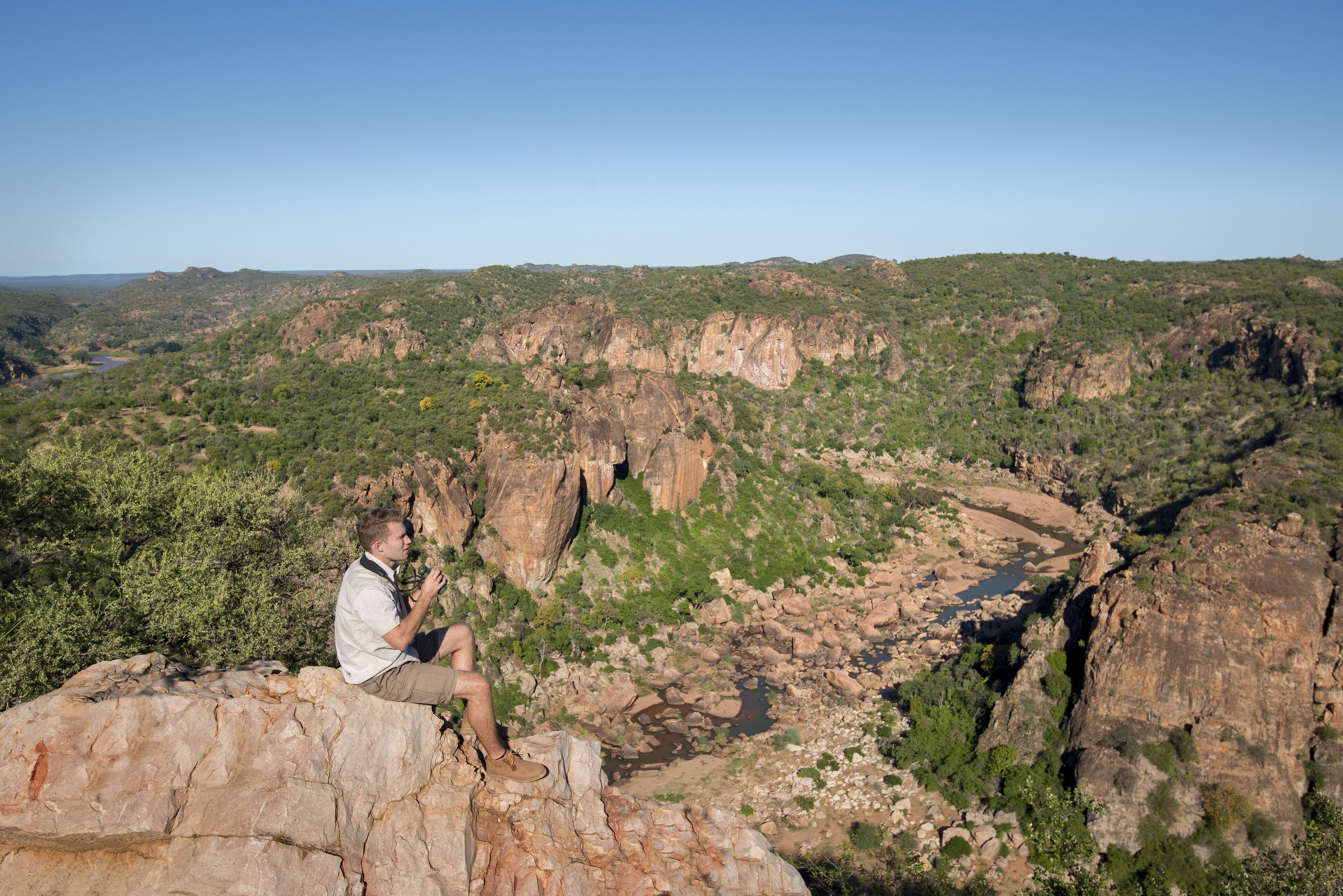 A person sitting on a rock overlooking a canyon with greenery and rocky terrain.