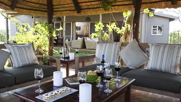 Un espacio de estar al aire libre con techo de paja, muebles de madera y una mesa dispuesta con copas de vino, una vela y pasteles.