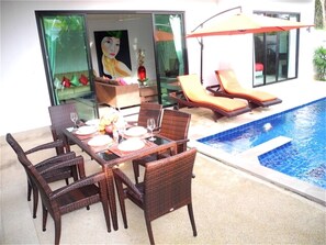 A dining area with wicker chairs and a table set for four, near a swimming pool with lounge chairs.