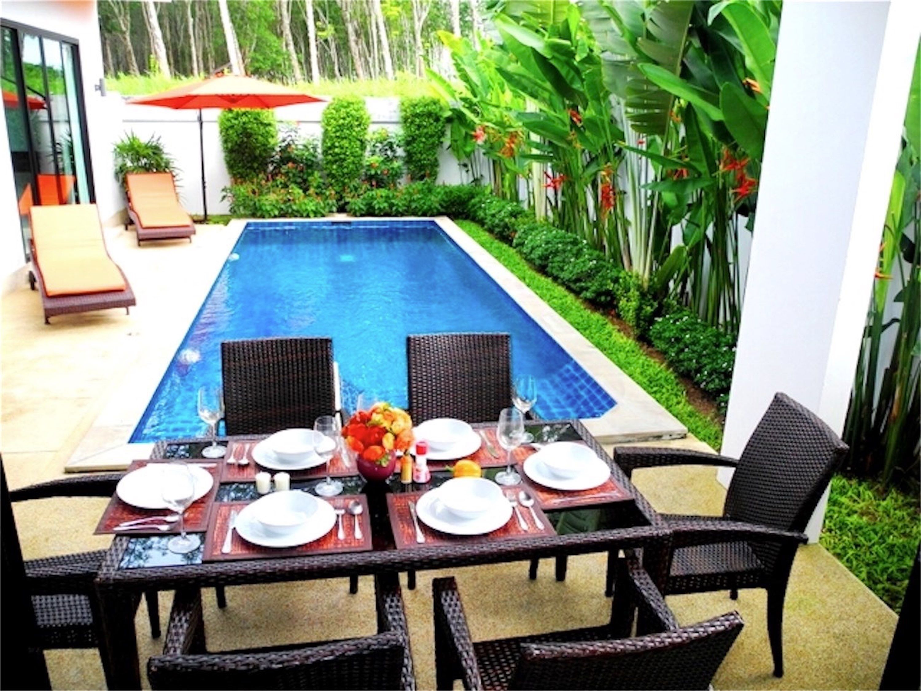 Uma área de refeições junto à piscina, com uma mesa posta para dois, rodeada por cadeiras de vime e um guarda-sol vermelho ao fundo.