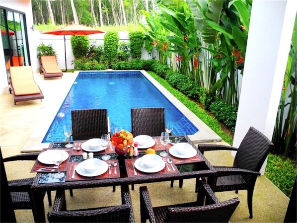 A poolside dining area with a table set for two, surrounded by wicker chairs, and a red umbrella in the background.